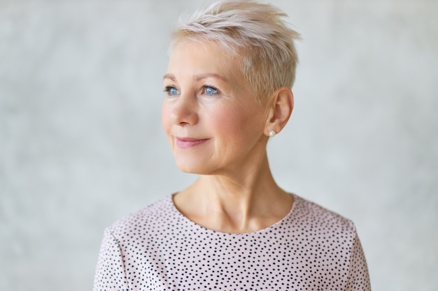 Close up beautiful attractive middle aged European lady with stylish haircut and neat make up looking away with confident smile posing isolated against marbled wall