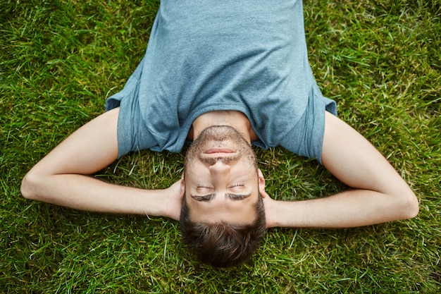 Close up of beautiful bearded mature caucasian male in blue t-shirt lying on grass in summer evening with closed eyes and relaxed face expression.