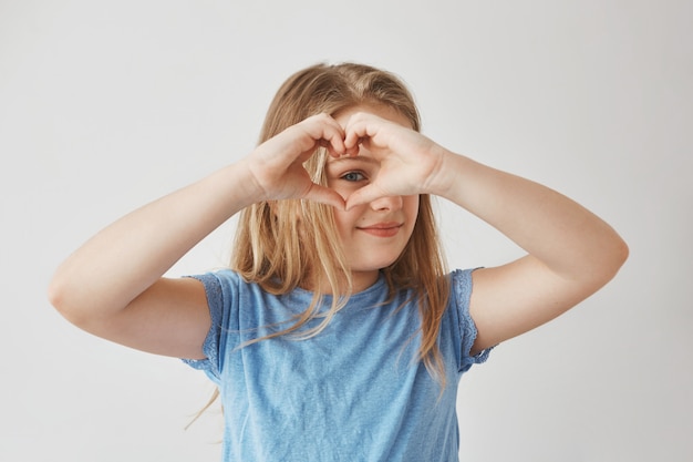 Close up of beautiful blonde girl making heart with hands, looking through it , posing for photo with smile and happy expression.