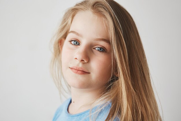 Close up of beautiful blonde girl with bright blue eyes holding head in three quarters, posing for photo with gentle smile.