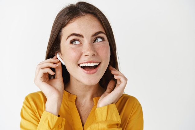 Close-up of beautiful caucasian woman listening music in wireless headphones, looking up and smiling, standing over white wall