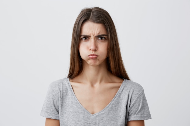 Free photo close up of beautiful charming funny young caucasian female with dark long hair in casual gray shirt inflate cheeks,  with unsatisfied and offended face expression, having fun on grad