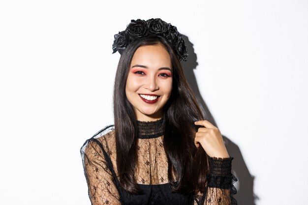 Close-up of beautiful elegant asian woman in black wreath and gothic lace dress smiling, standing over white background, dressed-up for halloween party.