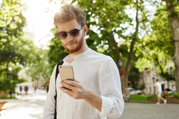 Close up of beautiful ginger male with beard and earring in stylish glasses texting with friend, going home after hard day on work.