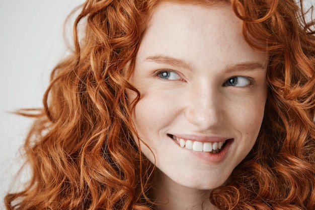 Close up of beautiful girl with curly red hair and freckles smiling biting lip .