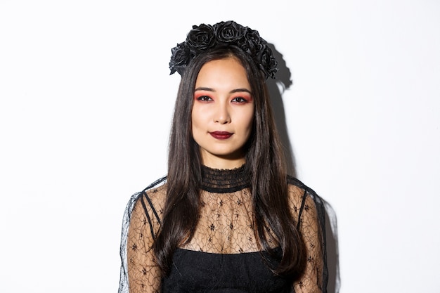 Free photo close-up of beautiful gothic girl with black wreath, getting dressed for halloween party, standing over white background in wicked witch costume.