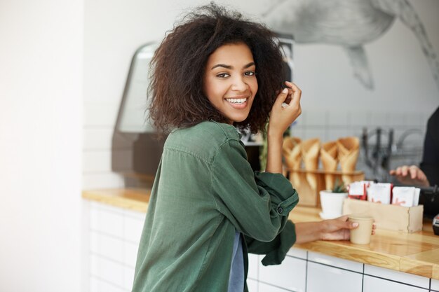 Close up of beautiful joyful african student woman with dark wavy hair in green cardigan sitting in cafe, drinking cup of coffee, smiling in camera. Woman waiting for her boyfriend after university.