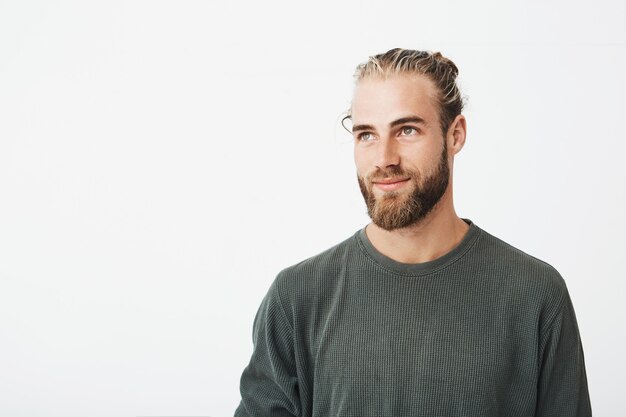 Close up of beautiful manly guy with light hair, fashionable hair and beard in grey shirt smiling and looking aside with pleasant expression.