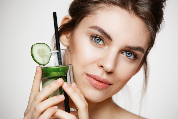 Close up of beautiful nude girl smiling looking at camera holding glass of water with cucumber slices over white background. Healthy nutrition. Beauty and skincare.