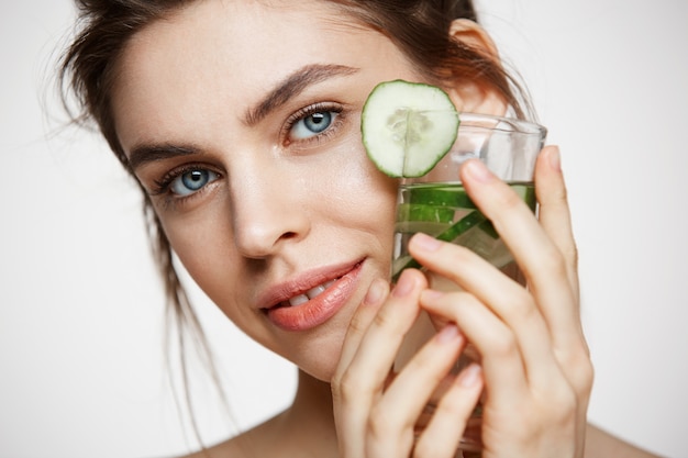 Close up of beautiful nude girl smiling looking at camera holding glass of water with cucumber slices over white background. Healthy nutrition. Beauty and skincare.