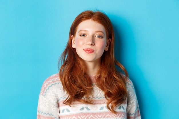 Close-up of beautiful redhead girl in winter sweater smiling at camera, looking satisfied and self-assured, standing over blue background.