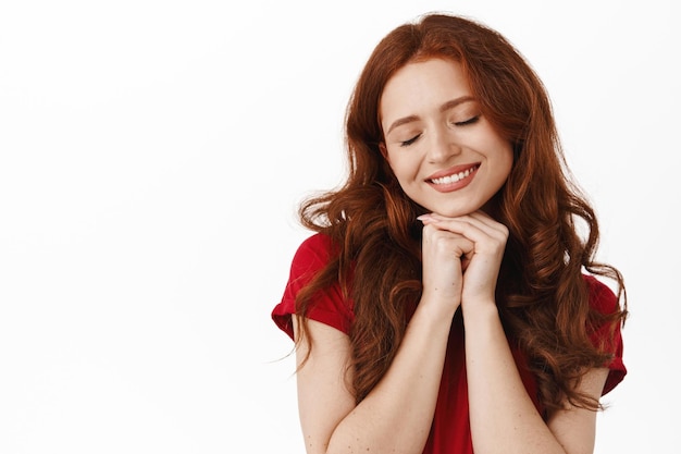 Close up of beautiful redhead woman with lovely white smile, feel romantic, daydreaming, thinking of something silly, close eyes, standing against white background