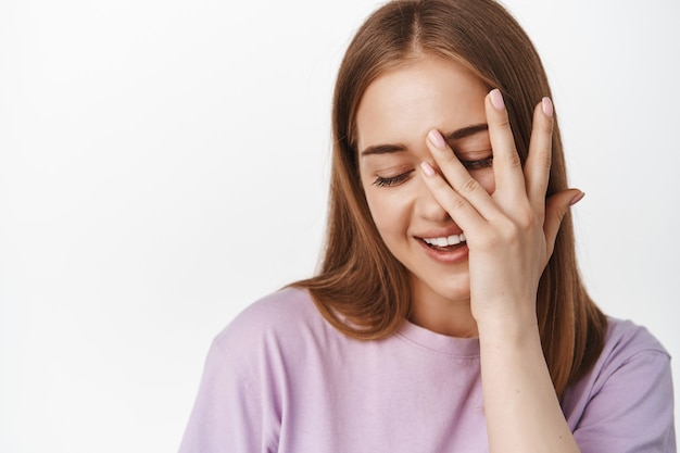 Close up of beautiful woman gently touch her facial skin, cover face with hand, look down coquettish and flirty, smiling, white wall