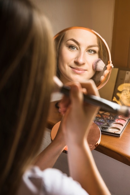 Close-up of a beautiful woman with hand mirror applying blusher on her face