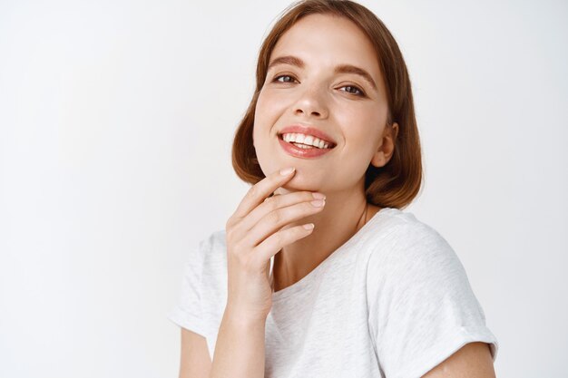 Close up of beautiful young woman with natural light make-up fresh face, touching chin and smiling tenderly, standing against white wall