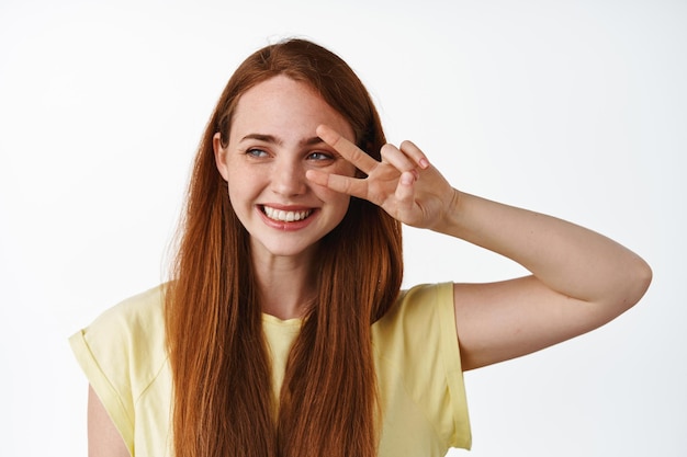Close up of beautiful young woman with natural pale skin and freckles, red long hair, show peace v-sign and smiling white teeth, standing against white background.