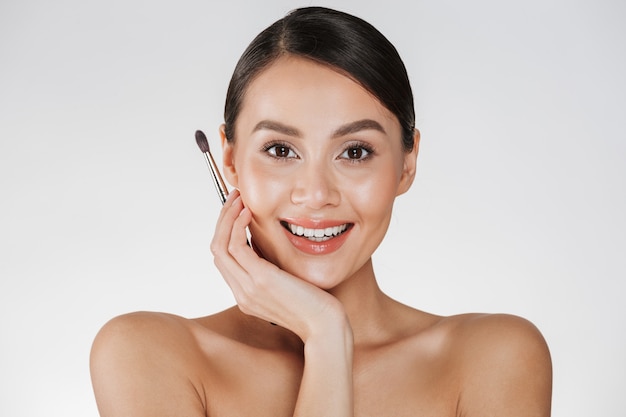 Close up beauty portrait of adorable brunette woman with soft skin holding makeup brush for eyebrows and looking on camera, isolated over white wall