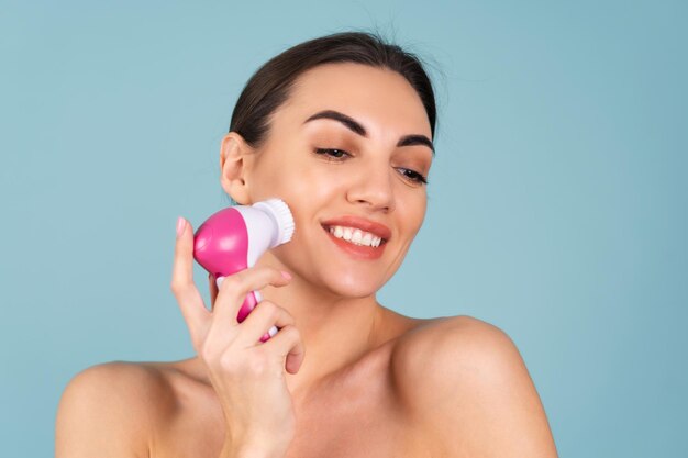 Close-up beauty portrait of topless woman with perfect skin and natural makeup, full nude lips, holding an electric facial brush. Facial cleansing with microvibrations