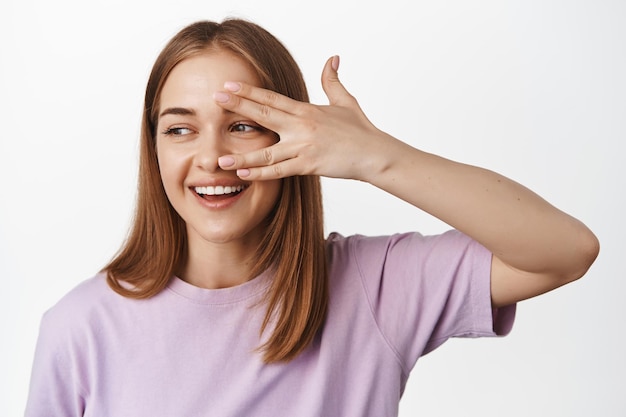 Close up of blond girl smiles, look through fingers aside, showing genuine happy and positive face expressions, beauty and skincare concept, white background