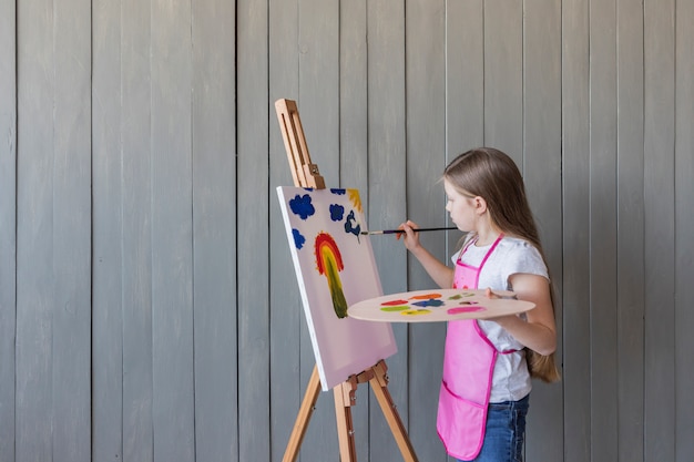 Close-up of a blonde girl painting with paint brush on easel standing against gray wooden wall