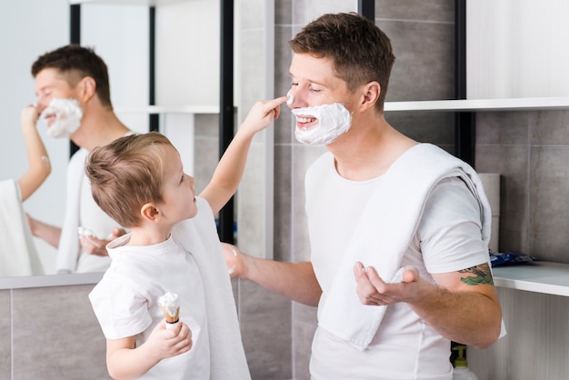 화장실에서 그의 아버지의 얼굴에 면도 거품을 적용하는 소년의 근접