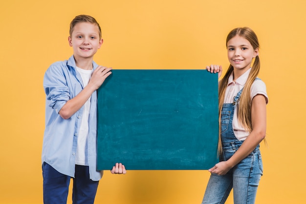 Close-up of a boy and girl holding green chalkboard against yellow backdrop