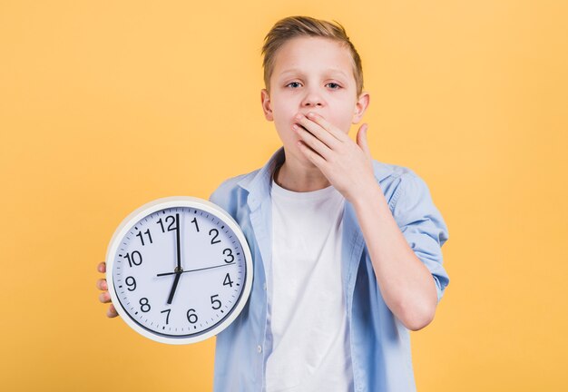 Close-up of a boy holding round white clock yawning with his hand on mouth standing against yellow background