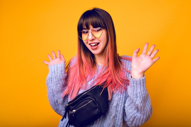 Закройте вверх по яркому позитивному портрету счастливой молодой женщины с розовыми волосами, вышедшими удивленными эмоциями, сердечными очками и уютным свитером.