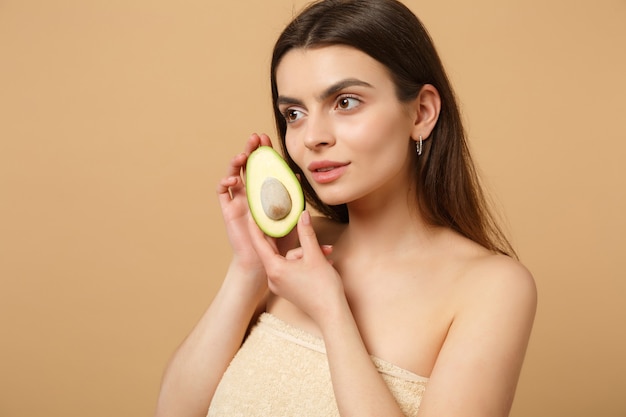 Close up brunette half naked woman with perfect skin nude make up holds avocado isolated on beige pastel wall