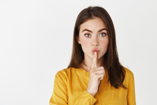 Close-up of brunette woman hushing, tell be quiet, share a secret and looking at front, showing taboo sign while standing on white wall