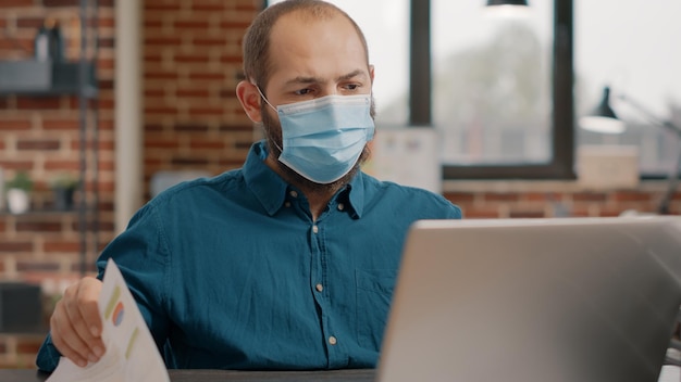 Close up of business man with face mask using laptop and documents on desk. Employee working with computer and charts papers to plan project and marketing strategy during pandemic.