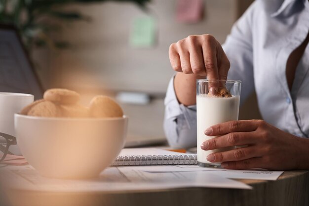 Close up of businesswoman eating breakfast at work and dipping cake in glass of milk