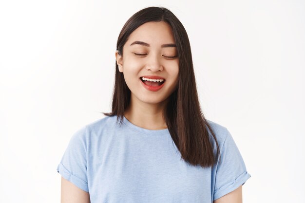 Close-up carefree positive relaxed charming asian happy girl close eyes smiling delighted singing express deep soul emotions shy look counting till see surprise gift stand white wall