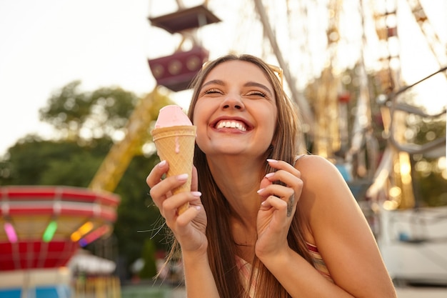 免费的特写照片迷人的年轻的黑发女士头上戴太阳镜,展示牙齿和微笑的幸福而在游乐园吃粉色冰淇淋蛋卷