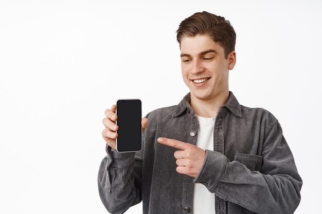 Close up of cheerful caucasian guy pointing finger at smartphone, showing mobile phone screen app, interface application, standing on white.