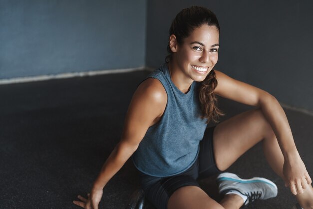 体育館の床に座っているクローズアップの陽気なフィットの格好良い若いブラジル人女性