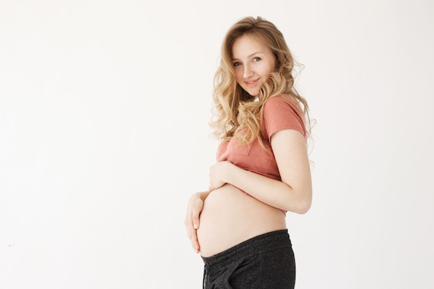 Close up of cheerful young good-looking pregnant mother with long blonde hair in casual clothes holding belly with hands