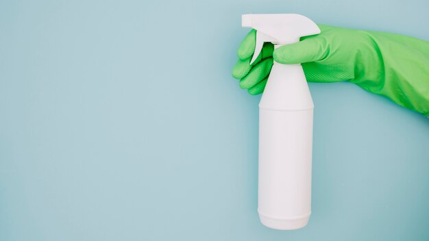 Close-up of cleaner's hand wearing green gloves holding white spray bottle on blue backdrop