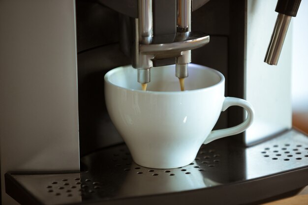 Close up of coffee machine pouring cappuccino, espresso, americano in white cup at home or cafe. Tasty and aroma hot drink. Food, nutrition, most popular drink for breakfest and break in worktime.