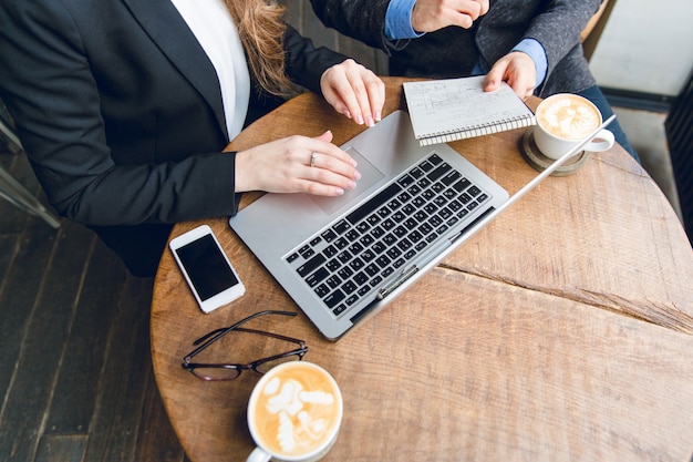 Close-up of a coffee table with two colleagues sitting, holding notebook and typing on laptop