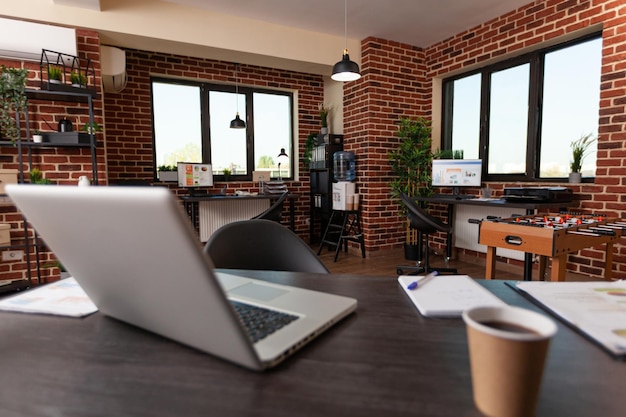 Close up of computer and office instruments on wooden table. Nobody in empty business space with laptop, cup of coffee and supplies used to work on startup company in boardroom.