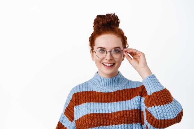 Close up of confident and determined young woman with ginger hair combed in hairbun wearing glasses smiling at camera standing against white background