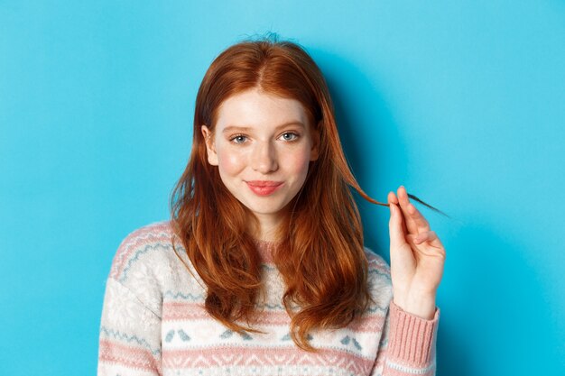 Close-up of confident and sassy redhead teen girl looking at camera pleased, playing with hair strand and smirking, standing over blue background