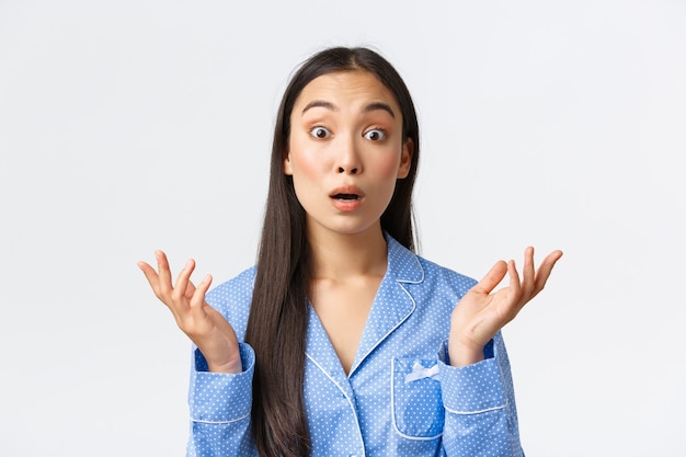 Close-up of confused and shocked asian girl in blue pajama stare startled and raising hands up, shrugging shoulders, cant understand what happened, standing unaware white background.