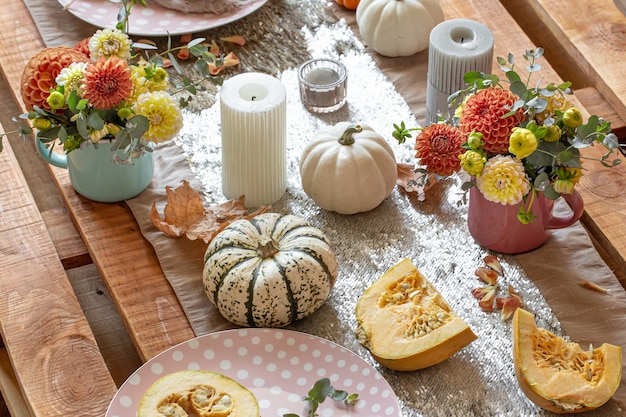 Крупный план уютных деталей декора праздничного осеннего обеденного стола с тыквами, цветами и свечами.
