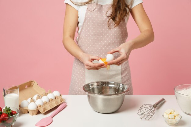 잘려진 여성 셰프는 앞치마 흰색 티셔츠를 입은 제과점이나 제과점을 닫고 스튜디오의 분홍색 파스텔 배경에 격리된 달걀을 들고 테이블에서 케이크나 컵케이크를 요리합니다. 복사 공간 음식 개념을 비웃습니다.