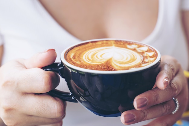 コーヒーショップカフェで女性の手にコーヒーカフェラテアートのカップのクローズアップ