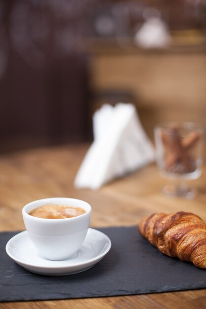 Close up of cup of coffee served with croissant in a cozy coffee shop. Coffee aroma.