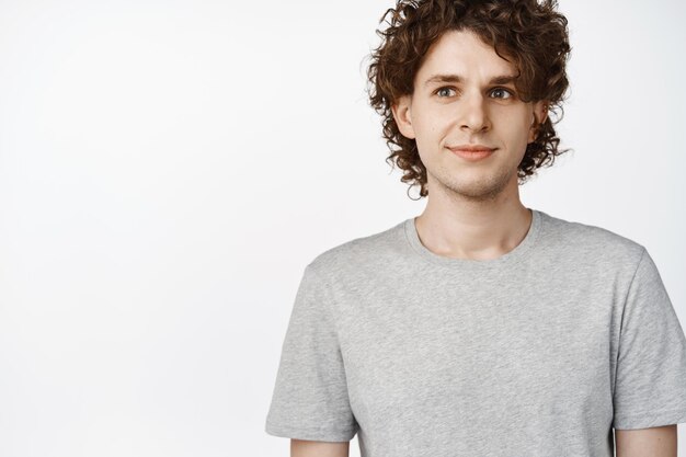 Close up of curly young man looking aside standing in gray tshirt over white background