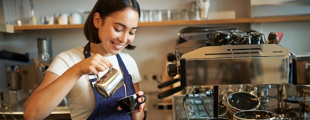 Free photo close up of cute asian barista girl making cappuccino doing latte art in cup with steamed milk